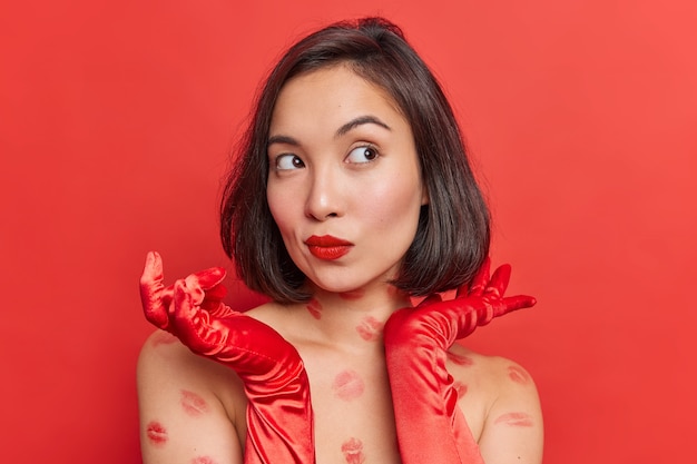 Superbe femme asiatique réfléchie aux cheveux noirs regarde pensivement loin porte d'élégants gants longs a des lèvres peintes en rouge des traces de baiser sur le corps pose des robes d'intérieur pour un rendez-vous romantique