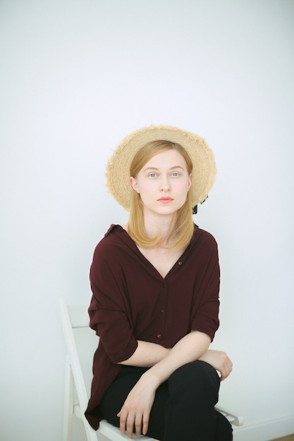 Superbe femme assise et regardant en chemise brune, pantalon noir et chapeau de soleil dans la chambre avec un fond blanc.