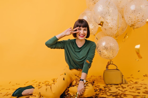 Superbe femme aux cheveux bruns posant avec signe de paix près de ballons de fête. Plan intérieur d'une fille heureuse en riant célébrant son anniversaire.