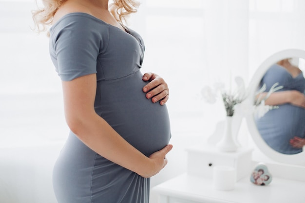 Superbe femme enceinte vêtue d'une longue robe grise posant dans un studio élégant embrassant son ventre et souriant à la caméra Jolie maman blonde attendant un enfant Doux intérieur du studio