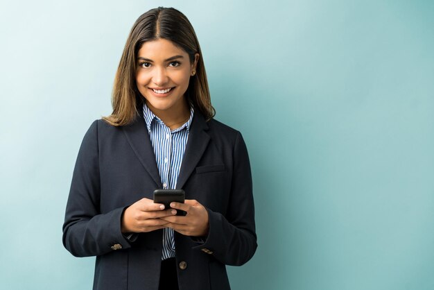 Superbe femme entrepreneure hispanique surfant sur un smartphone en se tenant debout et en établissant un contact visuel sur fond isolé