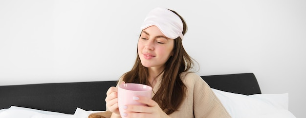 Superbe femme en pyjama et masque de sommeil tenant une tasse de café buvant du cappuccino dans son lit et