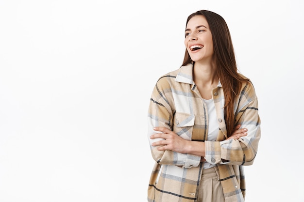 Superbe femme riant, regardant de côté le texte promotionnel de l'espace de copie avec un sourire heureux, debout en chemise à carreaux sur un mur blanc