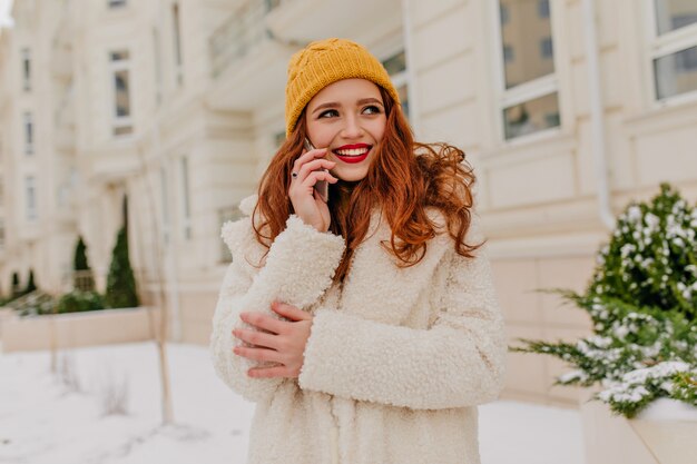 Superbe femme rousse souriante tout en posant avec le téléphone. Prise de vue en plein air d'une jolie femme au gingembre debout dans la rue le matin d'hiver.