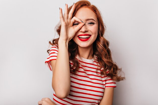 Superbe fille au gingembre avec une coiffure ondulée exprimant le bonheur. Intéressé jeune femme en tenue rayée s'amusant sur un mur blanc.