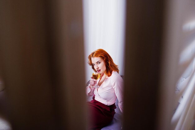 Superbe fille aux cheveux rouges en chemisier rose et jupe rouge avec un verre de vin à portée de main sur le lit de la chambre