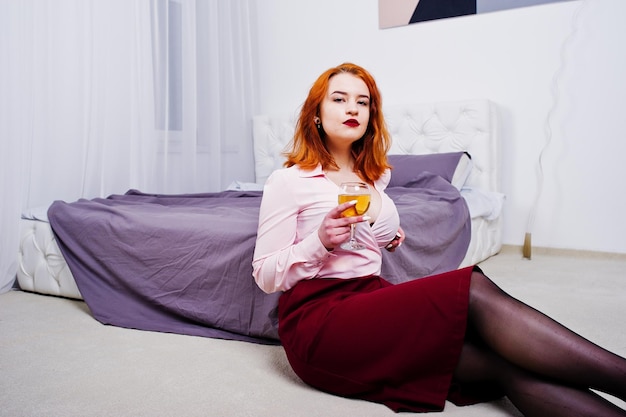 Superbe fille aux cheveux rouges en chemisier rose et jupe rouge avec un verre de vin à portée de main près du lit dans la chambre