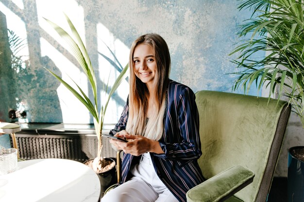 Superbe fille blanche assise dans un fauteuil avec le sourire. Blithesome dame caucasienne avec téléphone posant au café avec plaisir.