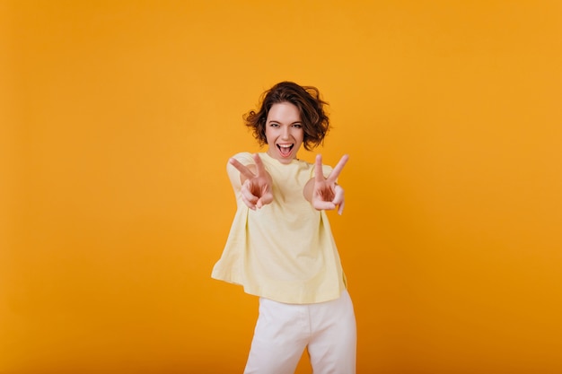 Superbe fille bouclée en t-shirt jaune danse drôle avec un sourire sincère. Femme aux cheveux bruns insouciante, froid et riant.