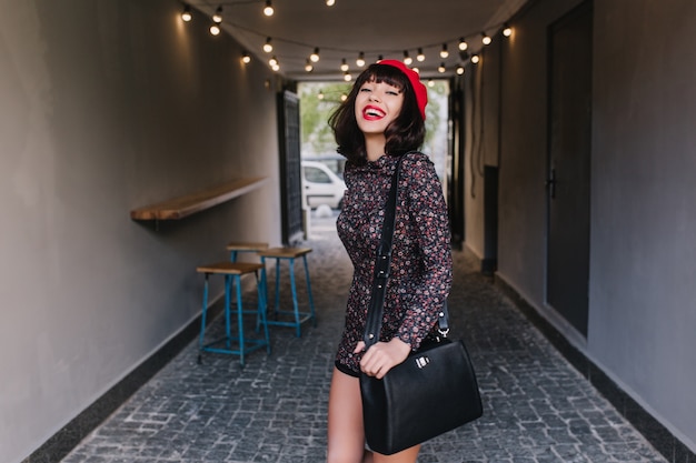 Superbe fille élégante aux cheveux courts foncés marchant après le travail portant un sac en cuir noir et souriant. Portrait d'adorable jeune femme brune portant des vêtements français vintage, s'amusant à l'intérieur