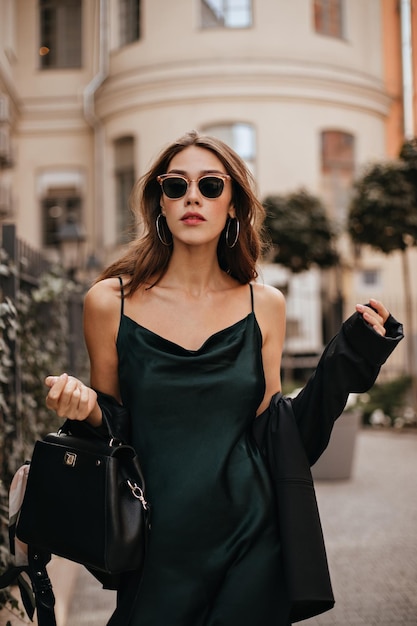 Superbe jeune brune aux lèvres rouges et tatouage sur le corps robe de soie verte sexy veste noire et sac lunettes de soleil à la mode debout à l'extérieur contre l'architecture de la vieille ville