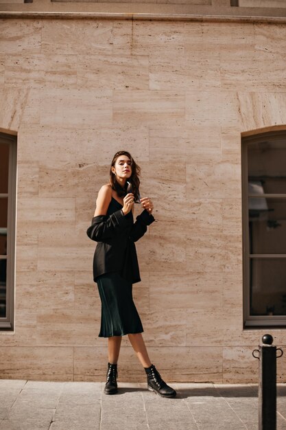 Superbe jeune brune en robe midi slip noir veste surdimensionnée et chaussures posant à l'extérieur sur fond de mur beige pendant la journée ensoleillée