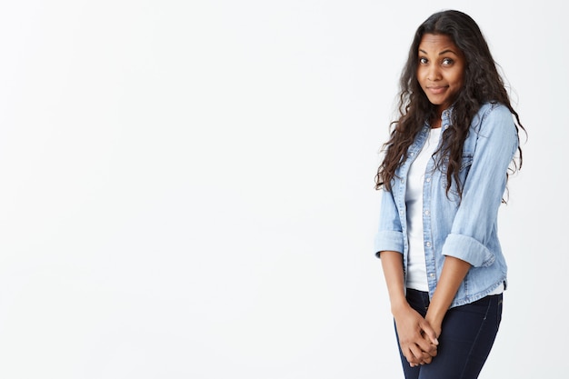 Photo gratuite superbe jeune femme afro-américaine avec une peau propre et sombre et un bel ensemble de fonctionnalités, vêtue d'une chemise bleu clair en jean sur un haut blanc, portant ses cheveux ondulés lâches