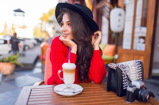 Superbe jeune femme au chapeau noir élégant et pull rouge vif assis dans un café en espace ouvert et boire du café avec du lait ou du cappuccino.