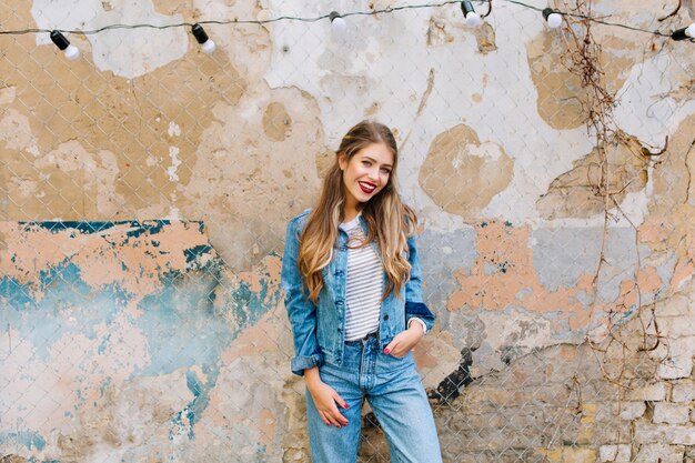 Superbe jeune femme blonde posant avec la main dans la poche isolée sur un fond grunge. Souriante jolie fille aux cheveux longs debout devant l'ancien bâtiment.