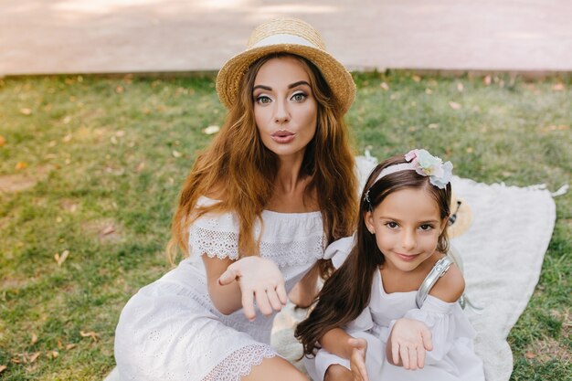 Superbe jeune femme et jolie fille partageant l'amour avec le monde posant pour la photo en pique-nique. Deux soeurs étonnantes dans des vêtements blancs similaires envoyant des baisers aériens se détendre sur l'herbe le matin.