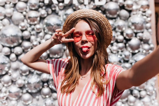 Superbe jeune femme en lunettes de soleil roses faisant selfie. Blithesome fille au chapeau de paille posant avec la langue près des boules disco.