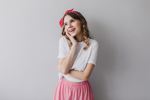 Superbe jeune femme avec ruban rouge pose rêveuse. Dame insouciante avec un sourire mignon debout.