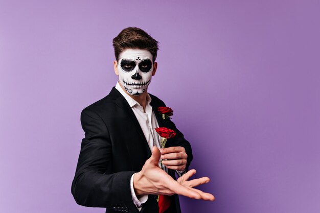 Superbe jeune homme avec l'art du visage en forme de crâne tenant une rose rouge dans ses mains, posant pour portrait sur fond isolé.