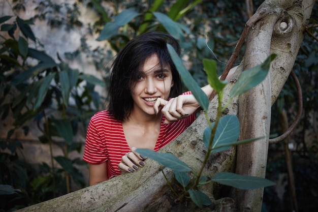 Superbe jolie fille regardant la caméra avec un sourire ludique, touchant les lèvres mystérieusement en se promenant dans le jardin ou le parc, entourée de plantes exotiques. Concept de nature, de personnes, de mode de vie et de loisirs