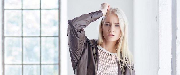 Superbe modèle féminin mince en robe blanche et veste noire posant sur une fenêtre lumineuse.