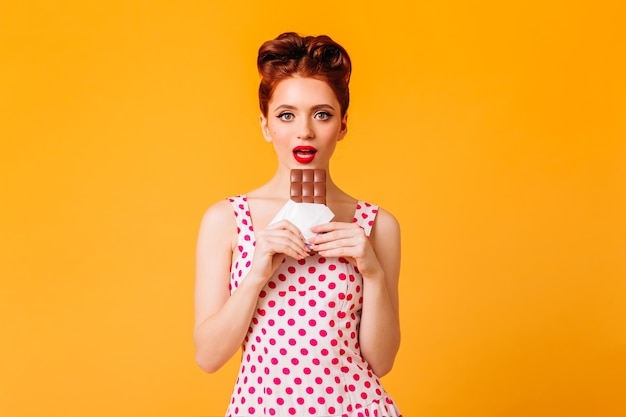 Superbe pin-up aux lèvres rouges mangeant du chocolat. Vue de face de la jeune femme au gingembre en robe à pois isolée sur l'espace jaune.