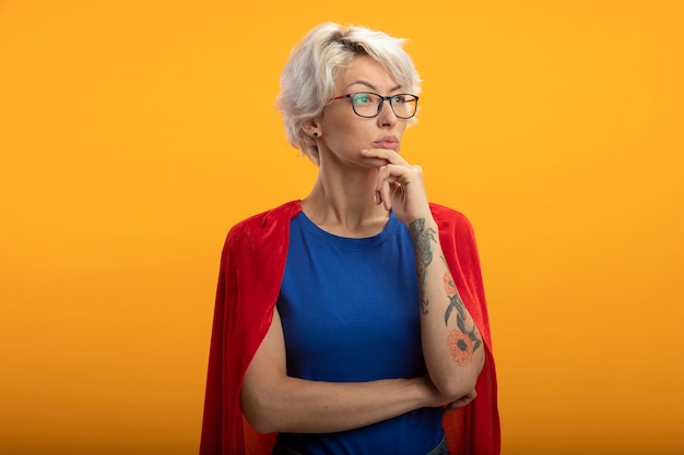 Superwoman confiant avec cape rouge dans des lunettes optiques met la main sur le menton et regarde à côté isolé sur mur orange
