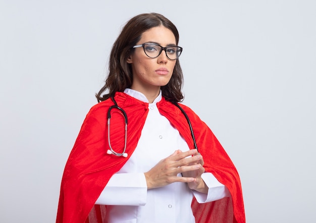 Photo gratuite superwoman confiant en uniforme de médecin avec cape rouge et stéthoscope à lunettes optiques tient les mains ensemble isolé sur mur blanc