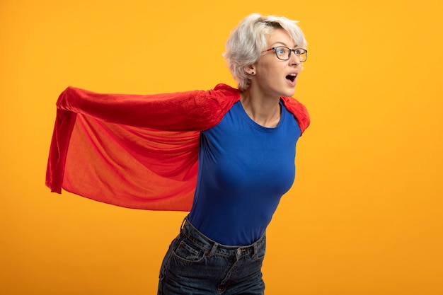 Photo gratuite superwoman impressionné dans des lunettes optiques détient cape rouge et regarde côté isolé sur mur orange