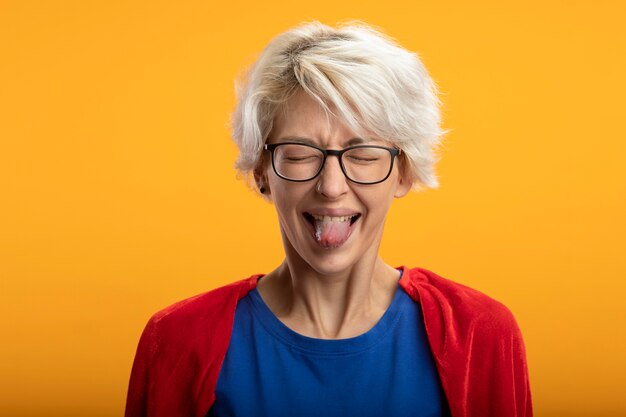 Superwoman mécontent avec cape rouge dans des lunettes optiques sort la langue isolée sur le mur orange