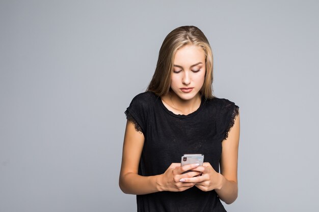 Surfer sur le net. Jolie jeune femme souriante et à l'aide de son téléphone intelligent en se tenant debout sur fond gris