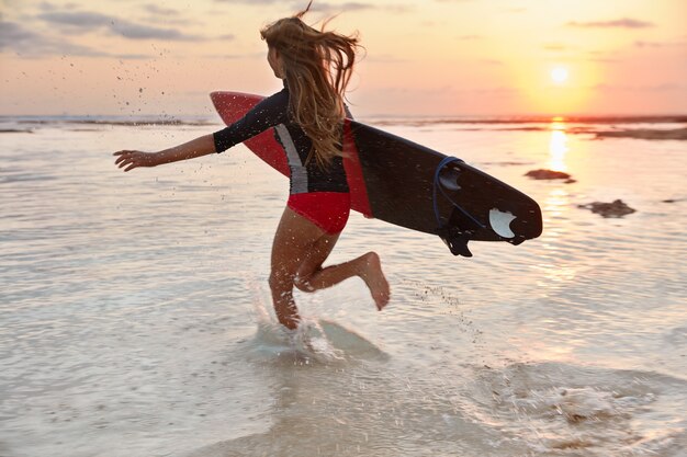 Un surfeur actif court dans l'océan avec bonheur, fait des éclaboussures d'eau, porte le bodyboard sous le bras