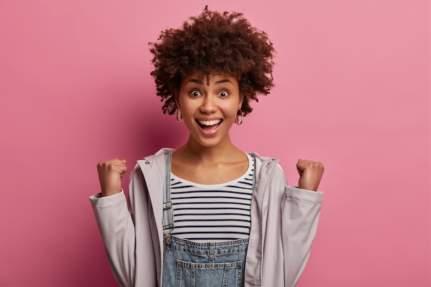 Surmotivante jeune femme triomphante avec une coiffure frisée, serre les poings et s'exclame de joie, a un large sourire, se réjouit du succès, porte un pull rayé, un anorak décontracté, pose sur un mur rose