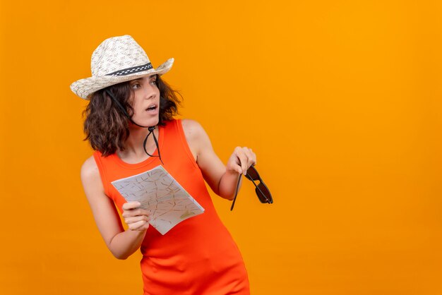 Une surprenante jeune femme aux cheveux courts dans une chemise orange portant un chapeau de soleil tenant une carte à la surprenante sur le côté