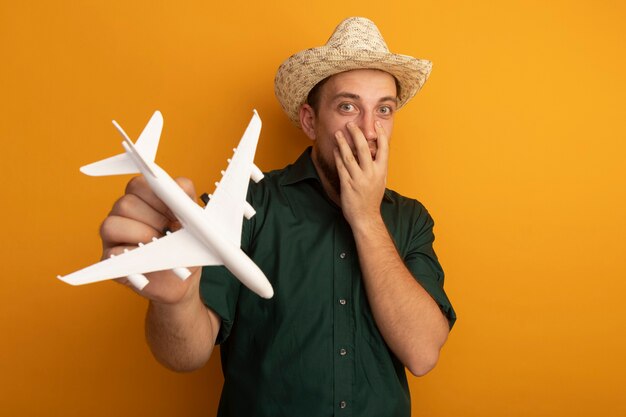 Photo gratuite surpris bel homme blond avec chapeau de plage met la main sur le visage et détient modèle avion isolé sur mur orange