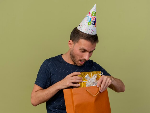 Surpris bel homme caucasien portant chapeau d'anniversaire tient et regarde la boîte-cadeau en papier sac à provisions isolé sur fond vert olive avec espace copie