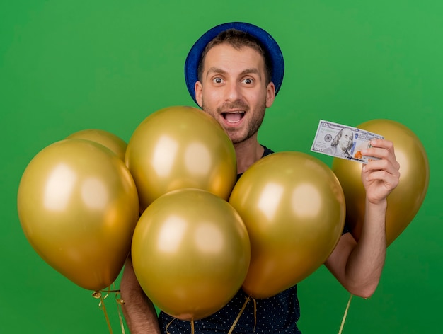 Surpris bel homme caucasien portant chapeau de fête bleu détient des ballons d'hélium et de l'argent isolé sur fond vert avec espace copie