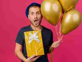 Photo gratuite surpris bel homme caucasien portant chapeau de fête bleu détient des ballons d'hélium et boîte-cadeau isolé sur fond rose avec espace copie