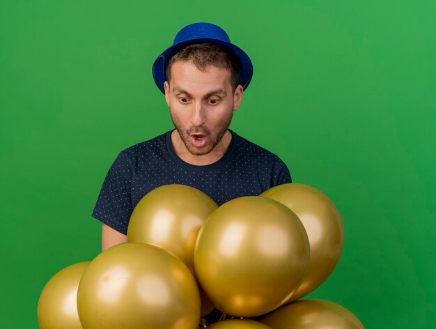 Surpris bel homme caucasien portant chapeau de fête bleu regarde des ballons d'hélium isolés sur fond vert avec espace copie