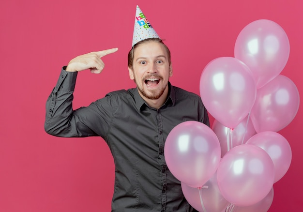 Surpris bel homme en chapeau d'anniversaire se dresse avec des ballons d'hélium pointant sur le cap