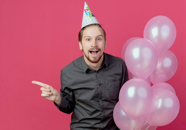 Surpris bel homme en chapeau d'anniversaire se dresse avec des ballons d'hélium pointant sur le côté