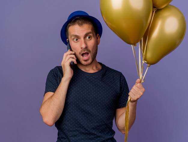 Surpris bel homme de race blanche portant un chapeau bleu détient des ballons d'hélium parlant au téléphone isolé sur fond violet avec espace de copie