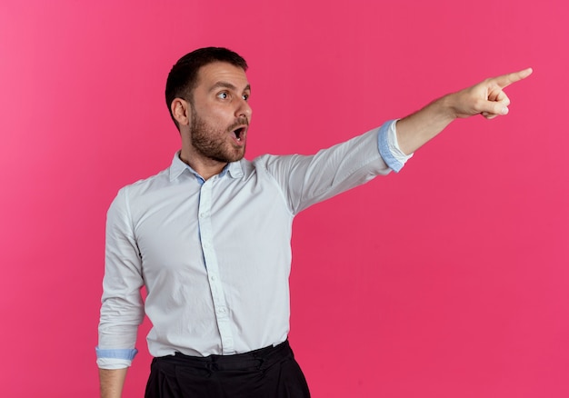 Surpris bel homme regarde et pointe sur le côté isolé sur mur rose