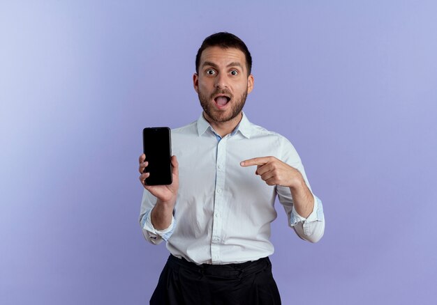 Surpris bel homme tient et pointe au téléphone isolé sur mur violet