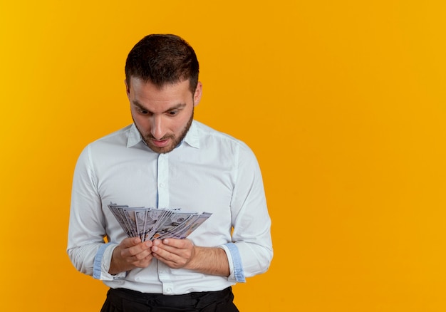 Surpris bel homme tient et regarde l'argent isolé sur un mur orange