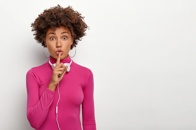 Surpris belle femme avec une coiffure afro, fait un geste de silence, porte des boucles d'oreilles rondes et un pull rose à col rond, écoute de la musique via un casque