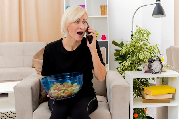 Surpris belle femme russe blonde est assise sur un fauteuil à parler au téléphone tenant un bol de chips à l'intérieur du salon