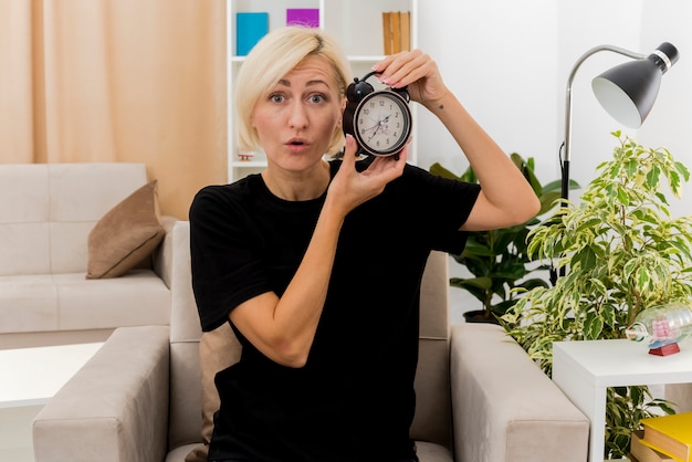 Surpris belle femme russe blonde est assise sur un fauteuil tenant un réveil à l'intérieur du salon