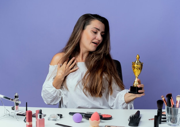 Photo gratuite surpris belle fille est assise à table avec des outils de maquillage met la main sur la poitrine et se penche sur la coupe du gagnant isolé sur le mur violet