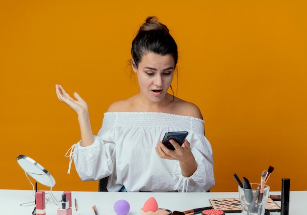 Surpris belle fille est assise à table avec des outils de maquillage et se penche sur le téléphone isolé sur le mur orange